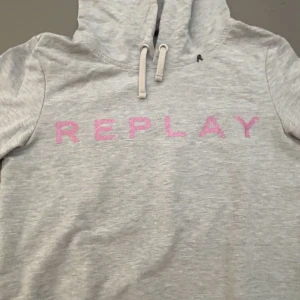Grå hoodie från Replay med rosa tryck - Snygg grå hoodie från Replay med stort rosa tryck över bröstet. Tröjan har huva med snörning och är tillverkad i mjuk bomull. Perfekt för chill dagar och streetstyle. Passar dig som gillar enkel men trendig design.