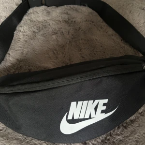 Svart Nike axelväska med logga och tunn träningsjacka. - Säljer en svart axelväska från Nike med klassisk vit logga framtill. Väskan har en justerbar rem och dragkedja upptill. Perfekt storlek för att bära med sig mobilen, plånbok och andra småsaker. Samt en tunn tränings jacka från Nike som kommer med. Perfekt set för träning. Storlek M på jackan.