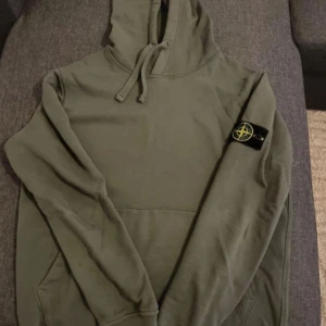 Olivgrön Stone Island hoodie - Säljer en olivgrön hoodie från Stone Island med klassisk patch på vänster ärm. Tröjan har huva med snörning och en stor magficka. Perfekt för dig som gillar streetwear och vill ha något stilrent med ikonisk logga.