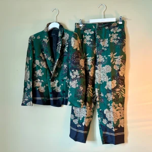 Grön blommig kostym från Zara Basic - Snygg kostym från Zara Basic med kavaj och byxor i grön och mörkblå färg, täckt av ett detaljerat blommigt mönster i vitt och beige. Kavajen har klassisk krage och knappar framtill, byxorna har raka ben och diskreta fickor. Materialet är glansigt och känns lyxigt. kavaj storlek M / Byxor storlek L