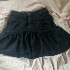 Svart volangkjol i jeans - Supersöt svart kortkjol i jeans med volang nertill och klassiska fickor fram. Kjolen har knapp och dragkedja i midjan, perfekt för en chill och trendig look. Passar till allt och ger en cool vibe.