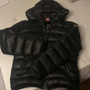 Svart Colmar pufferjacka med huva - Säljer en svart pufferjacka från Colmar med huva och två sidofickor med dragkedja. Jackan har ett stilrent, quiltat mönster och logotyp på ärmen. Perfekt för kalla dagar. Dragkedja framtill och mjukt, syntetmaterial. Lite av loggan på baksidan har lossnat men inget som syns.