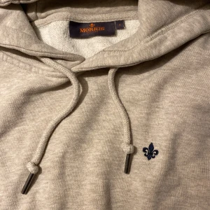 Beige hoodie från Morris - Snygg beige hoodie från Morris med mörkblå broderad logga på bröstet . Tröjan har huva med snörning och är tillverkad i mjuk bomullsmix som är perfekt för chill dagar. Passformen är normal och den har långa ärmar.