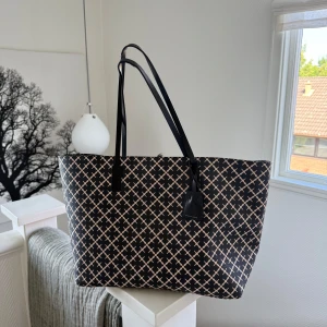 Svart shoppingväska By Malene Birger - Stor och rymlig shoppingväska från By Malene Birger med svart och beige rutigt mönster. Väskan har långa svarta axelremmar, innerfack med dragkedja och guldfärgad karbinhake. Perfekt för dig som vill ha plats för allt och samtidigt hålla stilen.