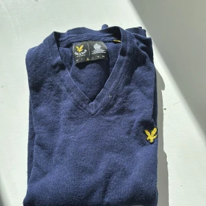 Marinblå v-ringad tröja Lyle & Scott - Snygg marinblå v-ringad tröja från Lyle & Scott med gul broderad logga på bröstet. Tröjan har långa ärmar och ribbade muddar vid ärmslut och nederkant. Perfekt för dig som gillar klassisk stil med en modern touch.