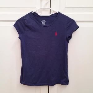 Marinblå t-shirt från Polo Ralph Lauren - Snygg marinblå t-shirt från Polo Ralph Lauren med klassisk röd logobrodyr på bröstet. Modellen är kortärmad och har rund hals. Tillverkad i mjuk bomull som är skön mot huden. Perfekt för dig som gillar stilrena och tidlösa plagg.