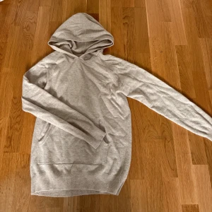 Grå-Beige stickad hoodie - Säljer en grå-beige kashmir hoodie med huva och långa ärmar. Tröjan har en stor magficka framtill och är mjuk och skön, perfekt för chill dagar. Enkel design som funkar till det mesta. 100% kashmir! 