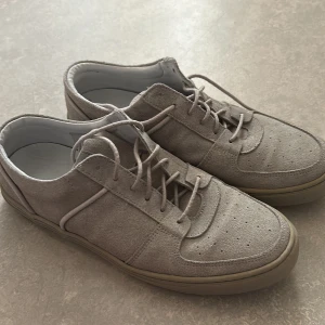 Beige mockasneakers från Jim Rickey - Säljer ett par snygga sneakers från Jim Rickey i beige mocka med rund tå och platt sula. Skorna har snörning och diskreta perforeringar på tån för extra stil. Perfekt för dig som gillar minimalistisk design och neutrala färger. Storlek 41 