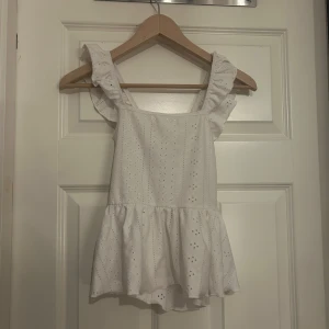 Vit blus med volang och knyt - Supersöt vit blus med broderade hålmönster, volangdetaljer på axelbanden och en luftig peplum-aktig nederdel. Blusen har knyt i ryggen för extra stilpoäng och är gjord i mjuk bomull. Perfekt för dig som gillar romantiska och trendiga vibes.