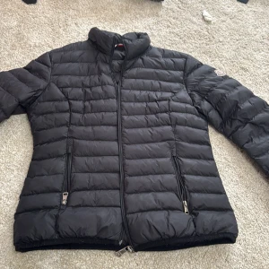 Svart dunjacka från Moncler - Snygg svart dunjacka från Moncler med horisontella quiltade paneler och två dragkedjeförsedda fickor. Jackan har hög krage, Moncler-logga på ärmen och stilren design. Perfekt för kalla dagar och enkel att matcha med din outfit.