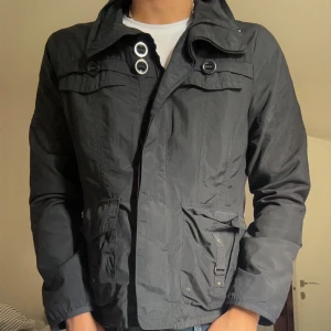 Field jacket  - Snygg svart vindjacka med stora fickor och metallringar vid kragen. 