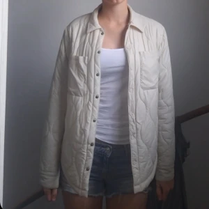 Quiltad beige jacka med knappar - Quiltad overshirt i beige med snygga tryckknappar framtill och två bröstfickor. Jackan har långärmad passform och rundad nederkant. Inte använd så ofta och det står att den är i storlek 170 men passar mig som har S