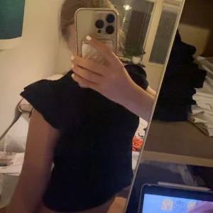 Svart topp med volangärmar - Säljer en svart topp med snygga volangdetaljer på axlarna. Modellen är figurnära och har korta ärmar med extra volym. Perfekt för dig som gillar en enkel men ändå cool look. Materialet känns mjukt och stretchigt mot huden.