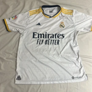 Real Madrid vit fotbollströja Adidas L - Snygg Real Madrid fotbollströja från Adidas i storlek L. Tröjan är vit med gula och mörkblå detaljer, har korta ärmar och klassiska Adidas-ränder på axlarna. Materialet är lätt och andas, perfekt för match eller träning. Tryck med Emirates Fly Better på bröstet.