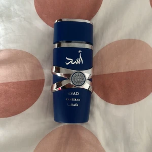 Lattafa Asad Zanzibar parfym - Snygg parfymflaska i matt mörkblått med silvriga detaljer och orientaliskt mönster på mitten. Flaskan har en unik form med silvriga band runt och text på både arabiska och engelska. Märket är Lattafa och doften heter Asad Zanzibar.
