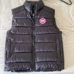 Canada Goose väst - Säljer en svart dunväst från Canada Goose med klassisk logga på bröstet. Västen är quiltad med horisontella sömmar, har hög krage och dragkedja framtill. Perfekt för lager-på-lager och håller dig varm under kyliga dagar.