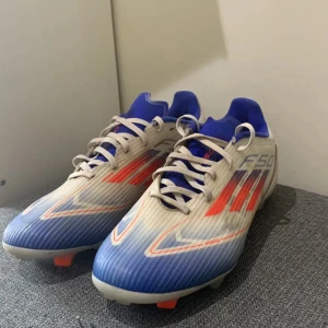 Adidas F50 blå och vita fotbollsskor - Säljer ett par Adidas F50 fotbollsskor med snyggt blått och vitt mönster, röda detaljer och klassiska tre ränder på sidan. Skorna har snörning, rund tå och platt sula med dobbar för gräsplan. Ovandelen är i syntetmaterial och insidan är blå. Perfekt för dig som vill sticka ut på planen.