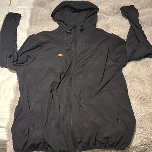 Svart vindjacka från Ellesse, M - Svart vindjacka från Ellesse med huva och dragsko, perfekt för blåsiga dagar. Jackan har hel dragkedja framtill, två sidofickor med dragkedja och Ellesse-logga på bröstet. Lätt och smidig design i syntetmaterial, passar dig som gillar sportig stil.