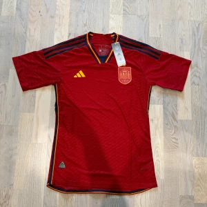 Spanien fotbollströja - Säljer en officiell Spanien fotbollströja från Adidas i röd färg med gula och marinblå detaljer. Tröjan har korta ärmar, V-ringad krage med röd, gul och marinblå rand. Materialet är lätt och andas, tillverkat med Parley Ocean Plastic. Slim fit-modell.