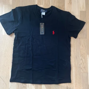 Svart t-shirt från Polo Ralph Lauren - Snygg svart t-shirt från Polo Ralph Lauren med klassisk röd broderad logga på bröstet. T-shirten har rund hals och korta ärmar. Perfekt basplagg med exklusiv känsla.