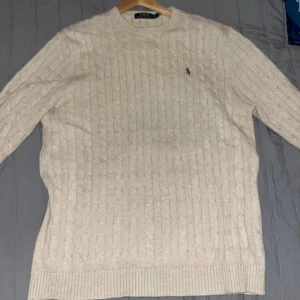 Beige kabelstickad tröja från Polo Ralph Lauren - Säljer en klassisk beige kabelstickad tröja från Polo Ralph Lauren med broderad logga på bröstet. Tröjan har rund halsringning och långa ärmar, perfekt för lager-på-lager-stil.