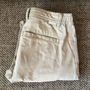 Beige chinos från Tom Tailor - Snygga beige chinos från Tom Tailor i slim fit-modell. Byxorna har klassiska sidofickor, bakficka och bälteshällor. Perfekta för en stilren och avslappnad look. Materialet är bomull med lite elastan för extra komfort.