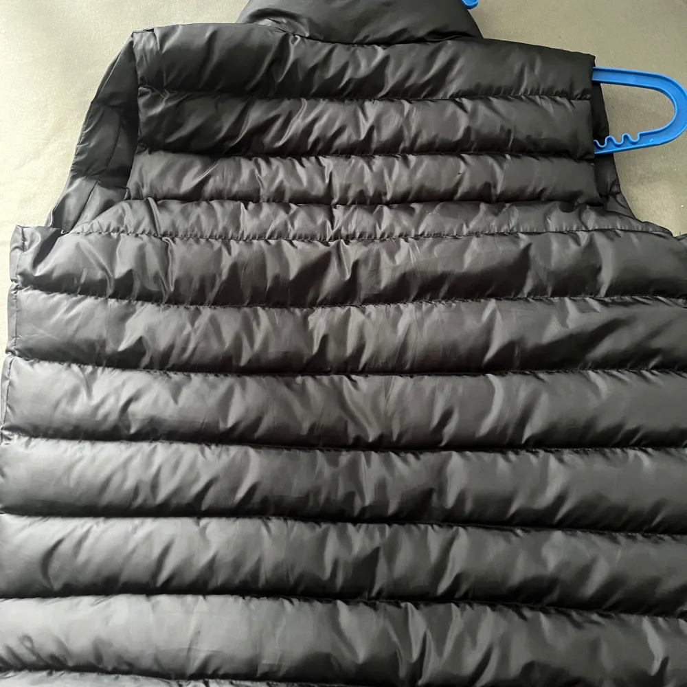 Säljer en svart dunväst från Moncler med hög krage och dragkedja framtill. Västen har två fickor med dragkedja och Moncler-logga på bröstet. Perfekt för lager-på-lager under kyliga dagar.. Takit.