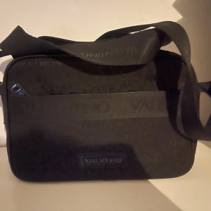 Svart axelväska från Valentino - Snygg svart axelväska från Valentino med diskret logomönster och bred axelrem. Väskan har dragkedja upptill och flera fack inuti för enkel organisering. Perfekt för att bära det viktigaste med stil.