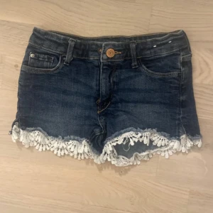 Mörkblå jeansshorts med spetskant - Snygga mörkblå jeansshorts med vit spetsdetalj längs bensluten. Klassisk femficksmodell med knapp och dragkedja framtill. Perfekta för sommaren och ger en söt touch till din outfit. För barn och är i storleken 110 och passar 4-5 åringar