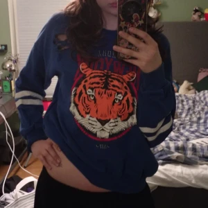 Blå sweatshirt med tigertryck från Gina Tricot - Säljer en blå sweatshirt från Gina Tricot med stort tigertryck och texten 'New York Bronx' framtill. Tröjan har långa ärmar med vita ränder och slitna detaljer på ärmar och bröst. Perfekt för en cool och avslappnad stil. Hör av dig vid frågor 💕