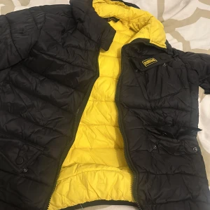 Svart och gul dunjacka från Barbour International - Säljer en svart dunjacka från Barbour International med gul insida. Jackan har dragkedja och flera fickor framtill. Perfekt för kyliga dagar och har en sportig look med Barbour-logga på bröstet.