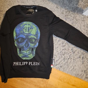 Philipp Plein - Svart sweatshirt från Philipp Plein med grön och blå "paljett" dödskalle tryck och Philipp plein text detalj på framsidan av tröjan. Har enbart körts i tvättmaskin 1 gång p.g.a allergi mot medlena i nya kläder, men den är Oanvänd och i nyskick ännu. Kommer i Storlek L/Large säljs för 1500 kr. Vid snabb affär 1000. Finns i Gränna att hämtas. OBS OBS OBS Vid frakt av tröjan står ni för porto kostnade själva 