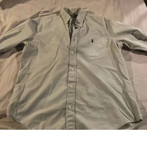 Grå skjorta från Polo Ralph Lauren - Klassisk grå skjorta från Polo Ralph Lauren i slim fit-modell. Skjortan har knappar framtill, button-down krage och den ikoniska broderade loggan på bröstet. Perfekt för en stilren look.