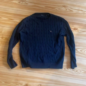 Mörkblå kabelstickad tröja från Gant - Säljer en mörkblå kabelstickad tröja från Gant med rund halsringning och diskret logga på bröstet. Tröjan har långa ärmar och ribbade muddar vid ärmslut och nederkant. Perfekt för en stilren och klassisk look.