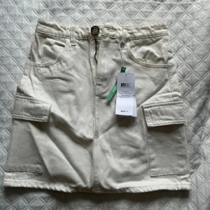 Beige kortkjol med fickor - Snygg beige kortkjol med praktiska fickor och dragkedja fram. Perfekt för en avslappnad stil. Kjolen har en knapp i midjan och är tillverkad i ett bekvämt material. Helt oanvänd!