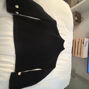 Svart ribbad stickad tröja med knappar - Säljer en svart ribbad stickad tröja med hög hals och långa ärmar. Tröjan har dekorativa silvriga knappar vid ärmsluten och en croppad passform. Perfekt för lager-på-lager och enkel att styla till olika outfits.