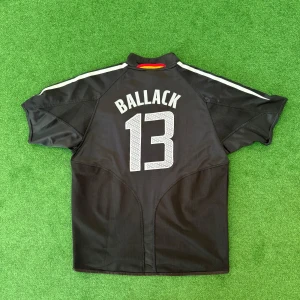 Tysklands fotbollströja Ballack 13 Adidas - Säljer en svart Tyskland fotbollströja från Adidas med Ballack och nummer 13 på ryggen. Tröjan har vita detaljer, tre stjärnor på bröstet och tysk flagga på ärmen. Klassisk Adidas-design med vita ränder på axlarna och inslag av gult och rött vid kragen.