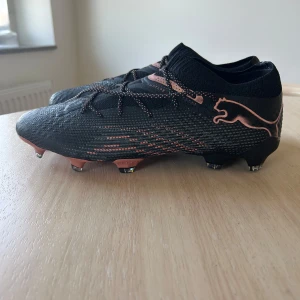 PUMA FUTURE 7 ULTIMATE LOW FG/AG ECLIPSE - SVART/KOPPARROS - Säljer ett par svarta fotbollsskor från Puma med snygga kopparfärgade detaljer och logga på sidan. Skorna har snörning och mönstrad ovansida för extra grepp. Nyskick, ej använda överhuvudtaget. Nypris 1349.