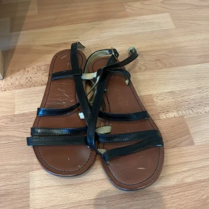 Svarta sandaler med remmar - Snygga svarta sandaler från Clarks med flera remmar och en platt sula. Perfekta för sommarens varma dagar. Sandalerna har en stilren design med en touch av guld på insidan av remmarna. 
