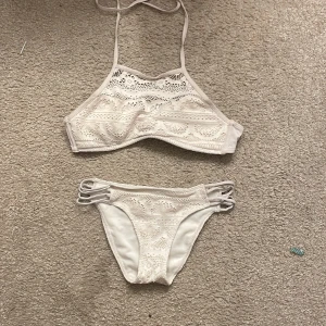 Vit spetsbikini - Snygg vit bikini med spetsdetaljer och smala axelband. Toppen har en hög hals och byxorna har dekorativa band på sidorna. Perfekt för en dag på stranden eller vid poolen.