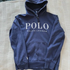 Marinblå hoodie från Polo Ralph Lauren - Säljer en marinblå hoodie från Polo Ralph Lauren med stor vit logga framtill och klassisk huva med dragsko. Tröjan har känguruficka och broderad logga på ärmen. Perfekt för en avslappnad och stilren look.
