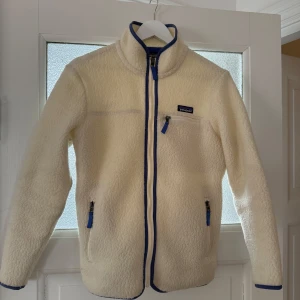Vit fleecejacka från Patagonia - Säljer en vit fleecejacka från Patagonia med blå detaljer. Jackan har dragkedja framtill och på fickorna, samt en hög krage. Lite nopprig under armarna men inga större defekter! 