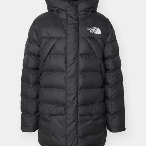 Svart dunjacka från The North Face - Säljer en svart dunjacka från The North Face med huva och logga på bröstet. Jackan är lång, har dragkedja och är perfekt för kalla dagar. Klassisk pufferstil med långa ärmar och stora fickor framtill.