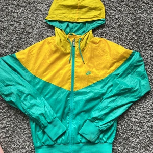 Nike vindjacka med huva i gult och turkost - Nike vindjacka i retrostil med huva. Jackan är tvåfärgad i gult och turkost, har dragkedja framtill och ribbade muddar. Perfekt för träning eller som en snygg streetwear-detalj. Klassisk Nike-logga på bröstet.