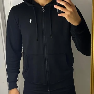 Svart hoodie från Ralph Lauren - Säljer en stilren svart hoodie från Ralph Lauren med dragkedja och justerbar huva. Den har en liten vit logotyp på bröstet och praktiska fickor framtill. Perfekt för en avslappnad look.