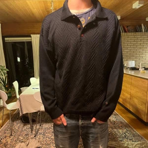 Mörkblå tröja från Bison Club - Säljer en mörkblå tröja från Bison Club med krage och knappar framtill. Tröjan har ett diskret mönster och långa ärmar. Perfekt för en stilren look.