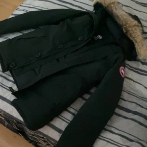 Äkta canada goose jacka  - Säljer en svart parkajacka från Canada Goose med avtagbar huva och pälsdetalj. Jackan har stora knappar framtill, sidfickor och klassisk logga på ärmen. Perfekt för kalla dagar.