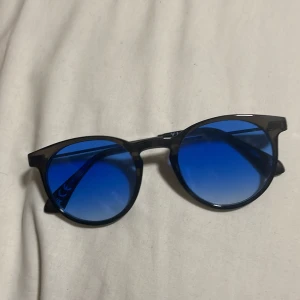 Blåtonade solglasögon från VILA - Snygga solglasögon med rundad form och blåtonade glas. Bågarna är svarta och tunna, vilket ger en stilren look. Perfekta för att sticka ut i sommarens soliga dagar.