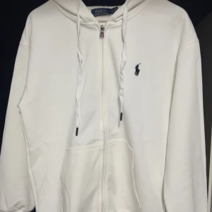 Vit hoodie från Polo Ralph Lauren - Säljer en klassisk vit hoodie från Polo Ralph Lauren med dragkedja och huva. Tröjan har broderad logga på bröstet och praktiska fickor framtill. Perfekt för en stilren och avslappnad look. Storlek S.