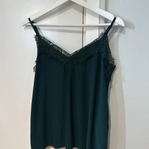 Mörkgrön linnetopp med spets från Amisu - Säljer en mörkgrön linnetopp från Amisu med tunna axelband och vacker spetsdetalj längs urringningen. Perfekt att styla till både jeans och kjol för en trendig look.